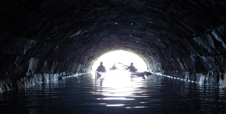 Bild. En tunnel. I öppningen några silhuetter av kajakare.