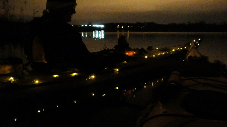Bild. En mörk bild. Christer i silhuett. Julgransbelysning på kajaken. Ån i bakgrunden.